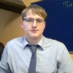 A profile of a man wearing a grey shirt, tie and glasses